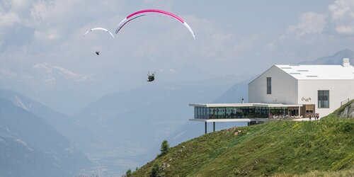 Zwei Gleitschirmflieger schweben über einer Bergwiese nahe einem modernen weißen Gebäude mit Terrasse. | © Harald Wisthaler